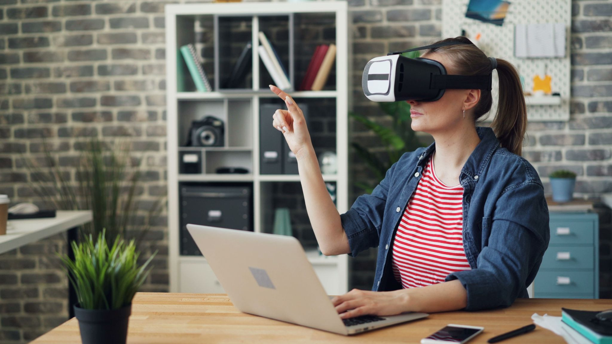 Person using a virtual reality headset and a grey laptop in their brick-walled home office