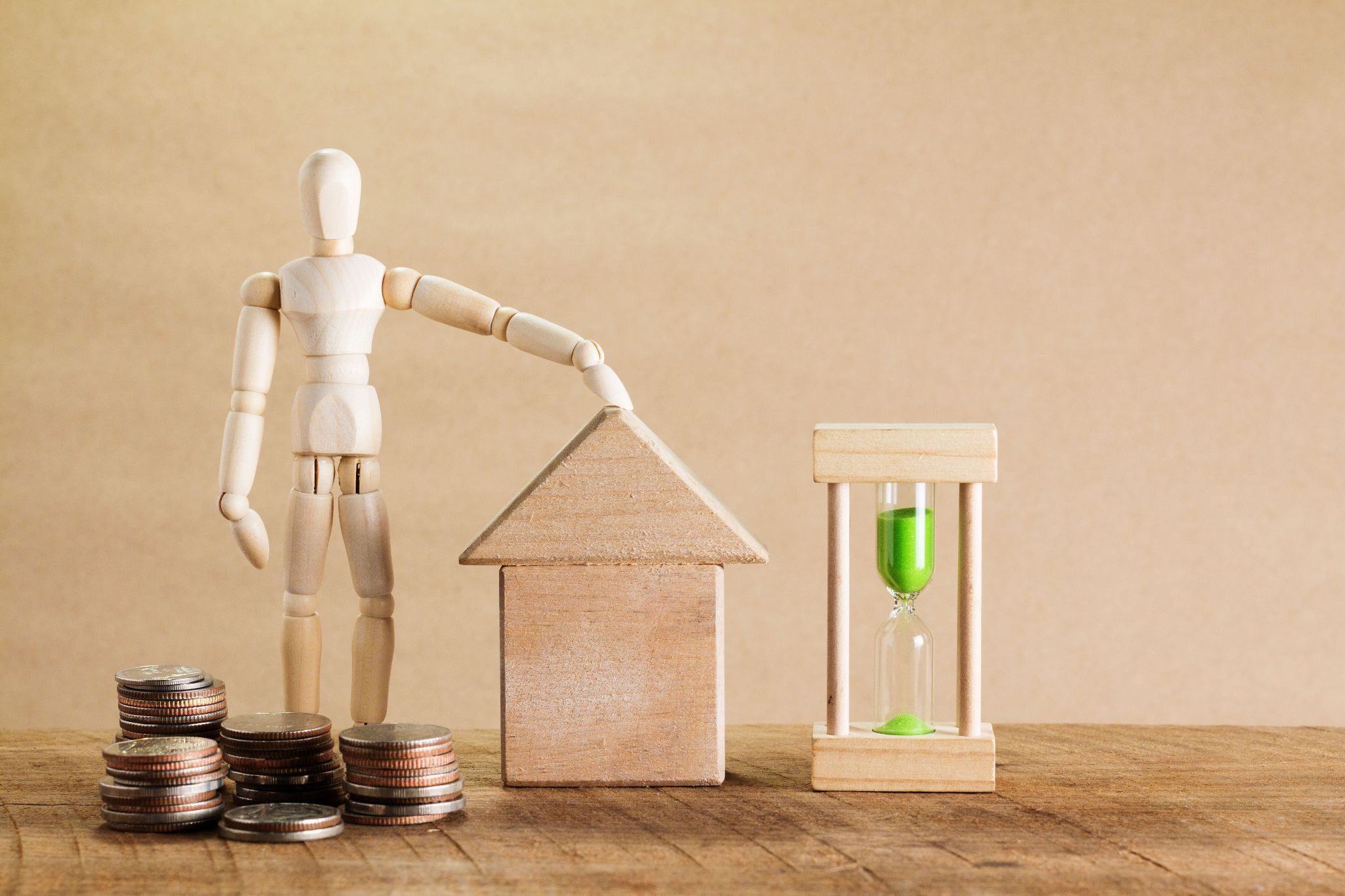 Small wooden mannequin facing the camera with its right hand o a wood model house next to a running hourglass